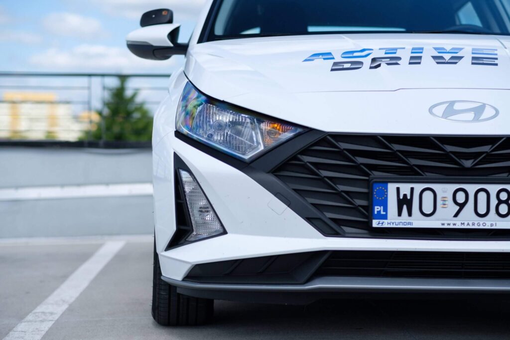 Zdjęcie to zbliżenie na przednią część samochodu Hyundai i20 do nauki jazdy, stojącego na parkingu. W kadrze widoczny jest prawy reflektor, atrapa chłodnicy z geometrycznym wzorem oraz zderzak. Na masce samochodu widoczne jest niebiesko-szare logo "ACTIVE DRIVE", które identyfikuje szkołę nauki jazdy.