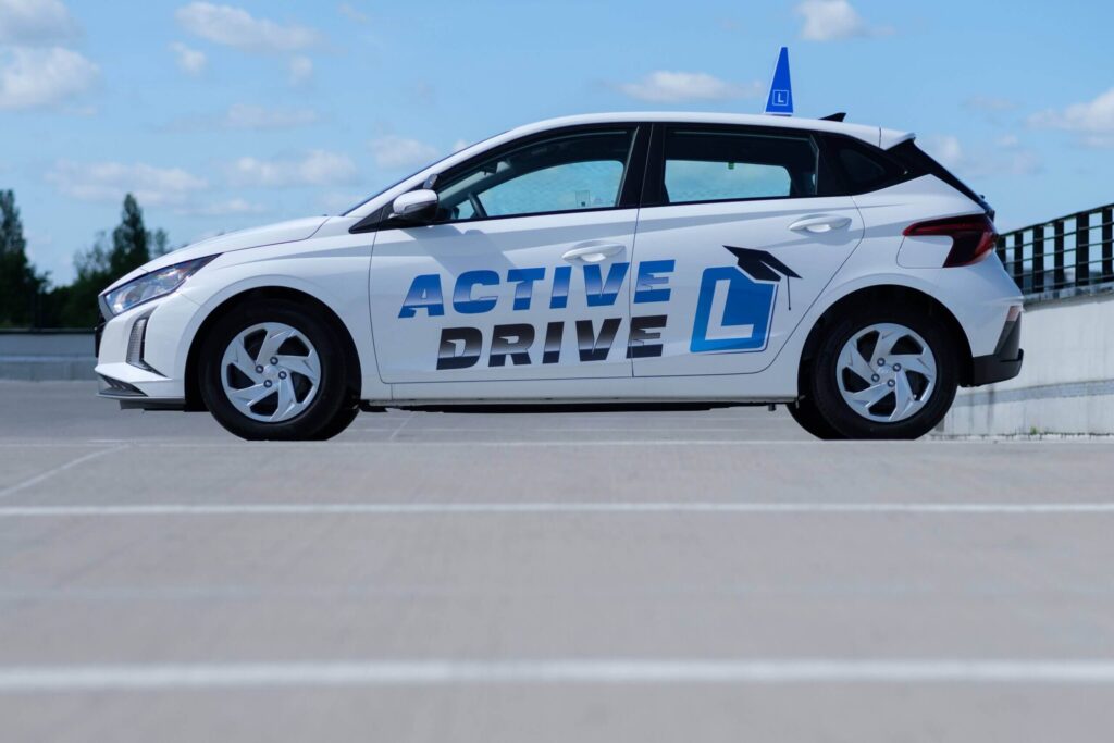 Zdjęcie przedstawia biały samochód Hyundai i20, który jest pojazdem do nauki jazdy. Samochód jest ujęty z boku i stoi na pustym, betonowym placu. Na drzwiach samochodu widoczne jest duże, niebiesko-szare logo szkoły "ACTIVE DRIVE", a obok niego duża litera "L" z biretem studenckim. Na dachu samochodu znajduje się również mniejsza, niebieska tabliczka z literą "L" w kształcie stożka.