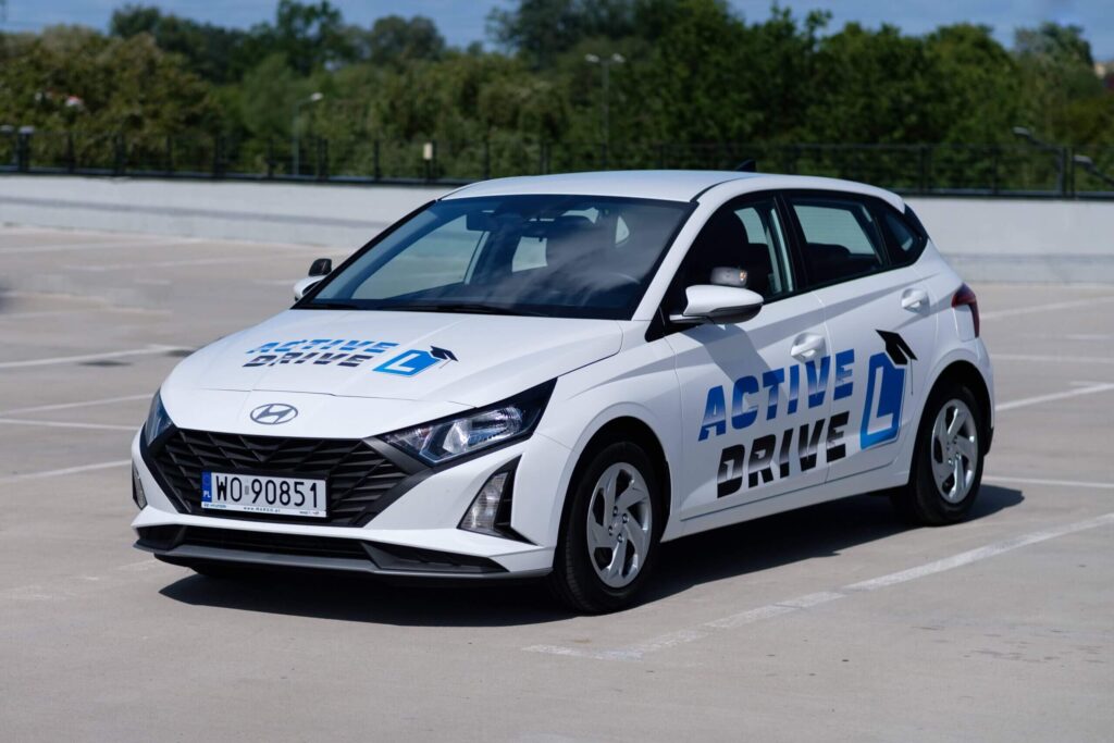 Zdjęcie przedstawia biały samochód osobowy marki Hyundai i20, zaparkowany na pustym parkingu pod gołym niebem. Jest to nowoczesny model, o sportowej, dynamicznej sylwetce. Na karoserii, na masce oraz na drzwiach bocznych, widoczne jest logo szkoły nauki jazdy z napisem "ACTIVE DRIVE" oraz charakterystyczna litera "L", symbolizująca samochód do nauki jazdy.