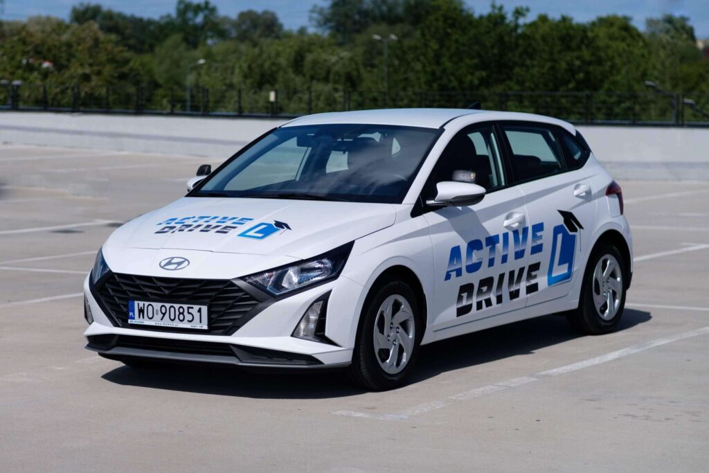Na zdjęciu widoczny jest biały samochód marki Hyundai zaparkowany na zewnętrznym parkingu. Na drzwiach i masce pojazdu znajdują się niebiesko-czarne napisy „ACTIVE DRIVE” oraz duży symbol „L”, co sugeruje, że jest to auto szkoleniowe dla kursantów. Tło zdjęcia obejmuje betonową barierę, drzewa i bezchmurne niebo.