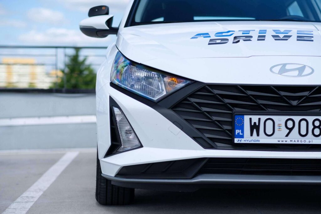 Na zdjęciu widoczny jest biały samochód marki Hyundai zaparkowany na parkingu. Na masce częściowo widoczny jest napis „ACTIVE DRIVE”, co sugeruje, że auto jest do nauki jazdy. Tablica rejestracyjna jest z oznaczeniem „PL”.