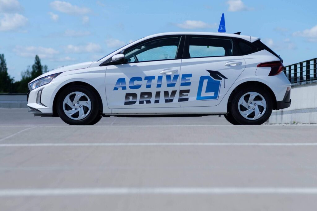 Na zdjęciu znajduje się biały samochód z dużym napisem „ACTIVE DRIVE” na bocznej części nadwozia. Na dachu oraz boku pojazdu widnieje niebieski znak „L”, co wskazuje, że jest to auto przeznaczone do nauki jazdy. Samochód stoi na utwardzonym placu, prawdopodobnie w strefie szkoleniowej. W tle widoczne są drzewa i niebo z lekkim zachmurzeniem.
